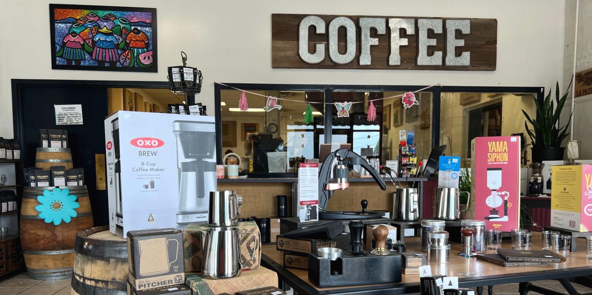 The interior of Columbia Street Roastery has a wooden sign that reads COFFEE along the back wall. Windows show a peek at the roastery, and the tables in the foreground offer a variety of coffee makers, pitchers, and books for sale.