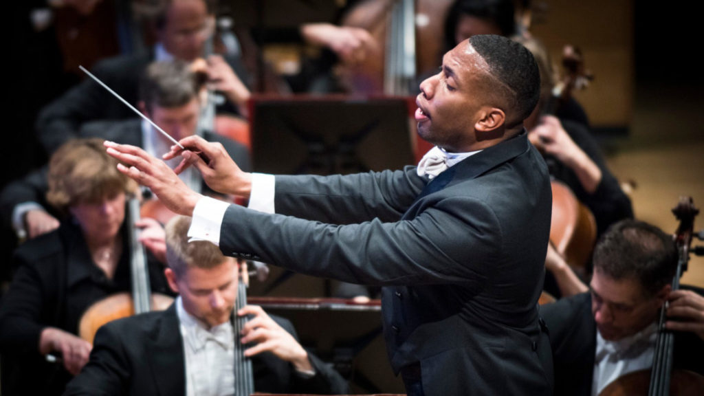 Photo of a Black man in a dark suit conducting an orchestra. Five other musicians playing cello are seen in the background.