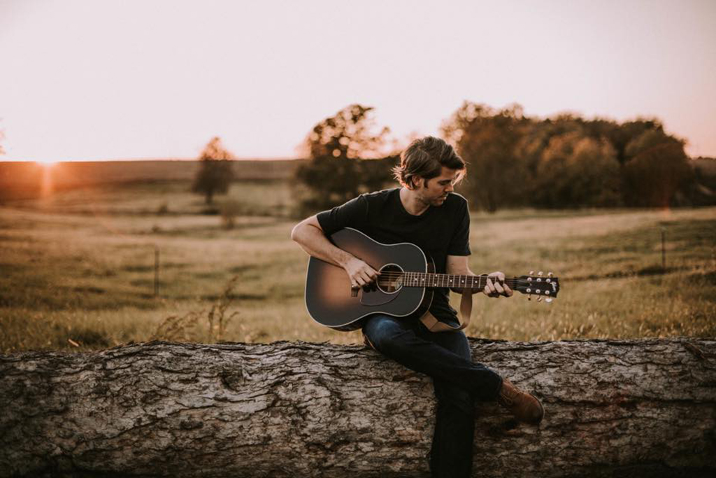 A man with short hair and a black t-shirt and jeans playing guitar while he sits on a log. In the distance you see a field with a few trees.