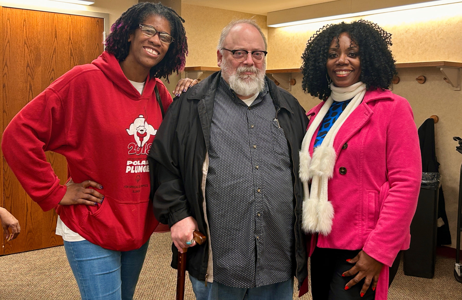 A Black woman in a red hoodie is on the left of a white man with a beard and glasses, and next to him on the right is a Black woman with a pink coat and white fluffy scarf.