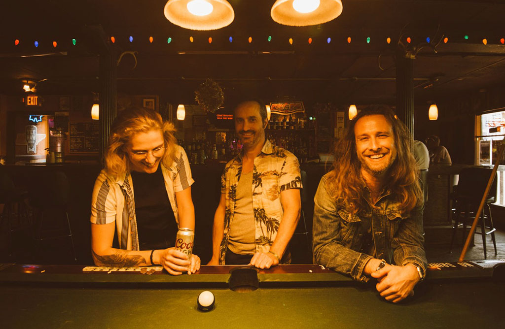 a casual and jovial moment among three individuals at a bar, standing around a pool table. The scene is cozy with warm lighting and string lights above, evoking a friendly and relaxed bar atmosphere. Two of them are smiling broadly, suggesting a moment of shared laughter or enjoyment, while the third person is leaning over the pool table, seemingly lining up a shot or preparing for a game.