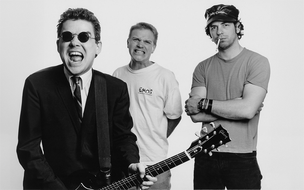 In the image, three men appear against a plain background, each exhibiting their own unique style and attitude. On the left, a man in a smart black suit and sunglasses exudes confidence with a wide, open-mouthed grin, gripping the neck of a guitar. At the center, another man, in a relaxed stance, wears a casual white t-shirt with the word "Emo's" printed on it, and he has a bemused or slightly aggressive expression. The third man on the right presents a rebellious pose, arms crossed with a cigarette dangling from his lips, sporting a cap, a simple tee, and a watch, projecting a too-cool-for-school vibe. Each member brings a different energy to the photo, suggesting a band with a diverse and dynamic character.