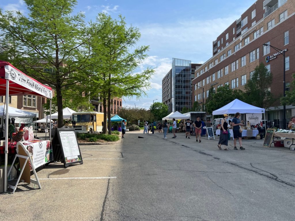 The outdoor farmers market in Parking Lot M of Downtown Champaign has several shoppers walking through the market of vendors.