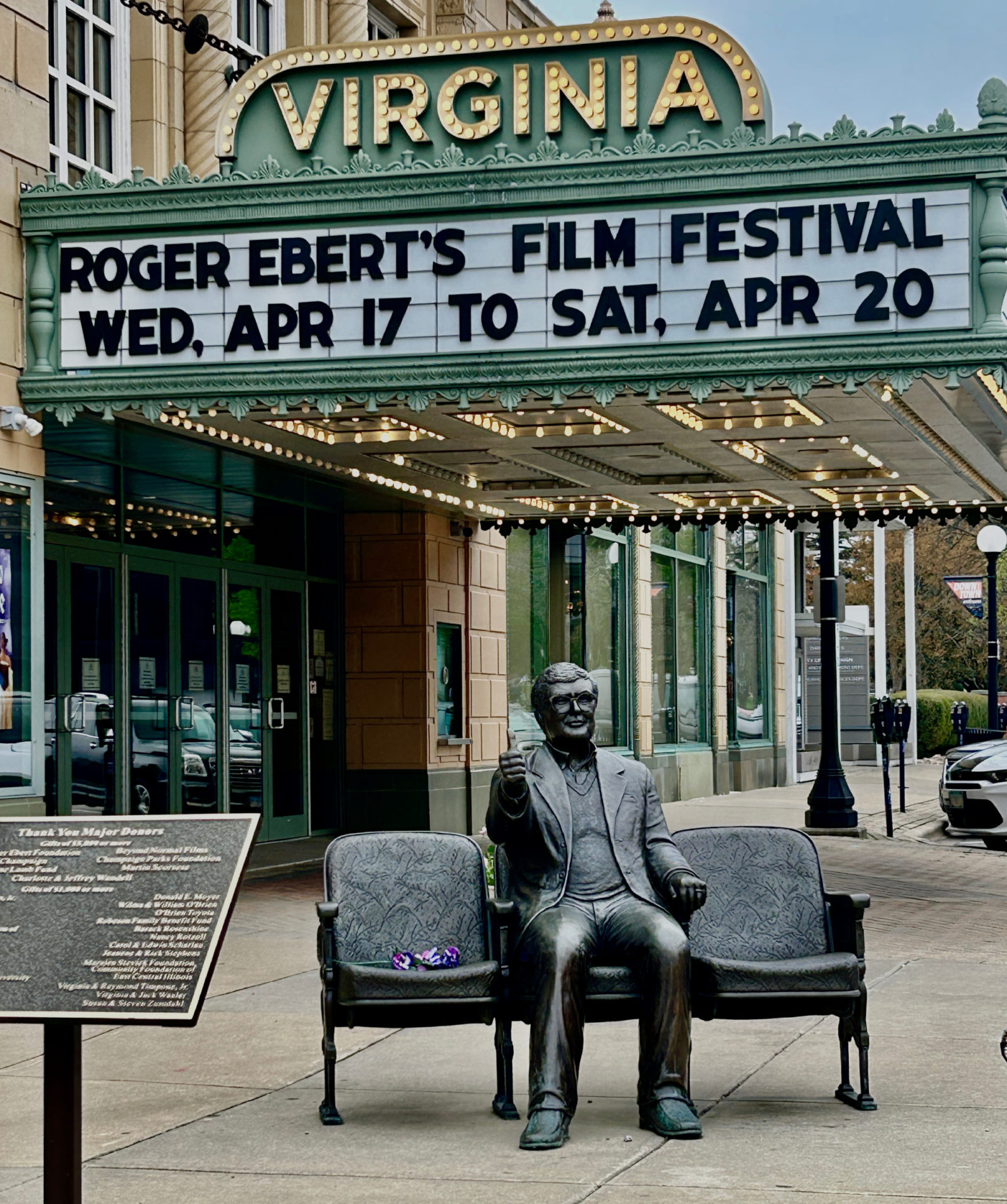 Photo of green film marquee with white background. The theatre is the Virginia. Text on the marquee reads Roger Ebert's Film Festival Wednesday April 17 to Saturday, April 20. A bronze statue of a man seated with his thumbs up is in front of the theatre on the sidewalk.