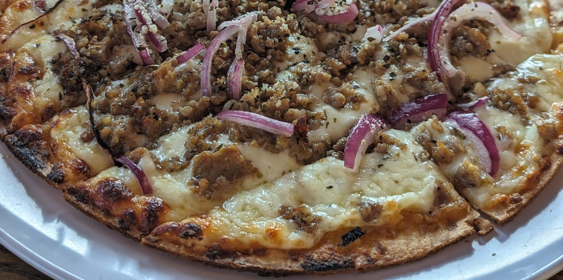 A flat pizza on a white plate with melty cheese covered in small pieces of sausage and slices of red onion.