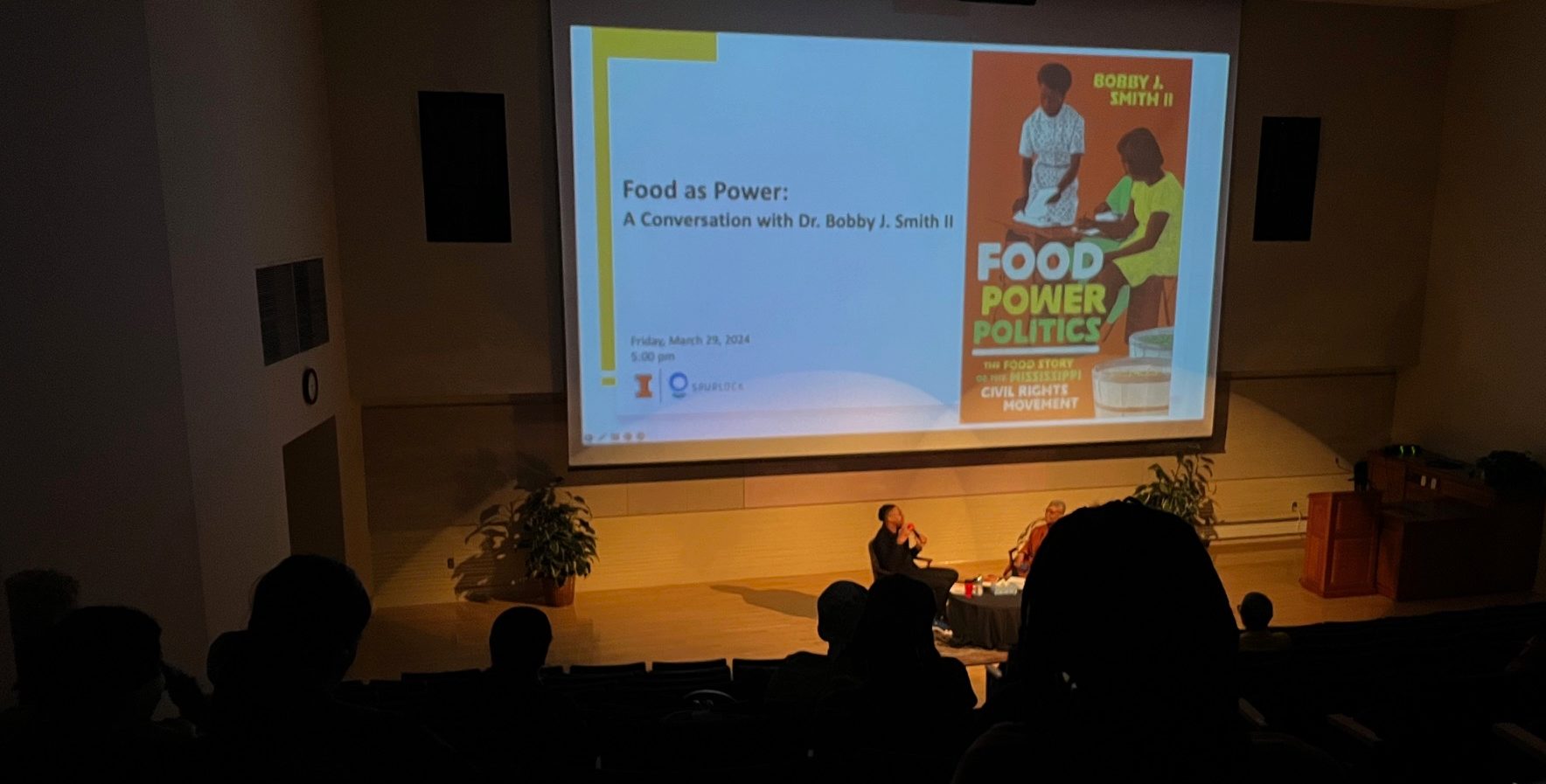 A screen reads Food as Power event and author Dr. Bobby J Smith II sits on a chair beside a Spurlock moderator.