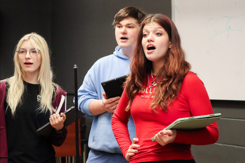 Photo of three white singers in rehearsal singing. One has long hair and a red shirt. The other two are blonde with glasses and one in the back wears a light blue hoodie.