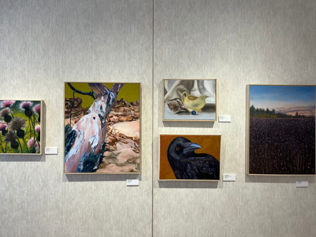 Five paintings are hanging on a white wall. They include landscapes with pine trees and blue sky; a yellow gravy boar and shell; a black raven's head, an abstract landscape; and pink blossoms on a canvas.