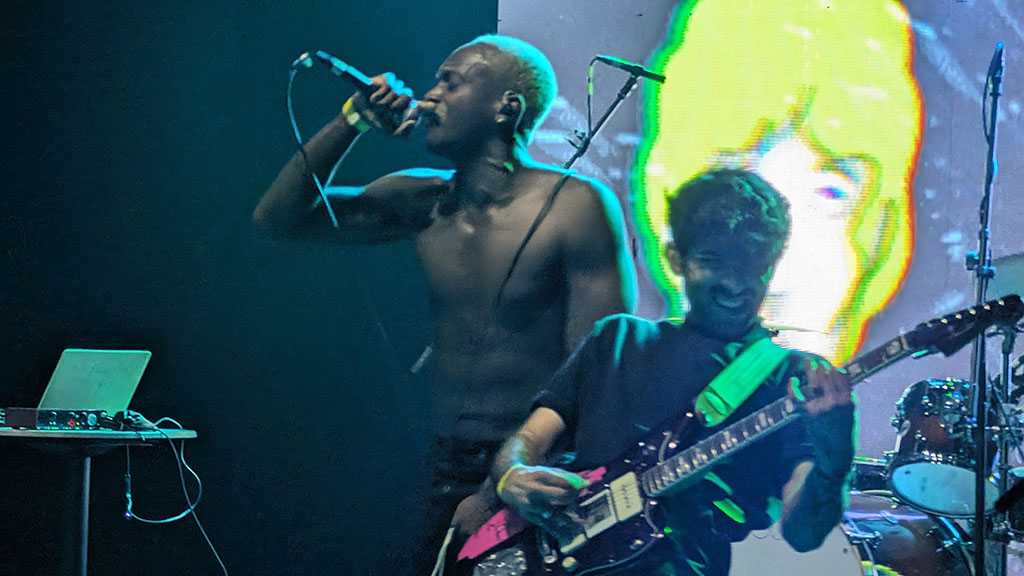 A vocalist and guitarist are performing together on stage, illuminated by the colorful display in the background. The vocalist, shirtless and using a handheld microphone, sings with an open mouth, while the guitarist, holding an electric guitar, is partly obscured by the microphone stand, looking towards the vocalist with a smile.