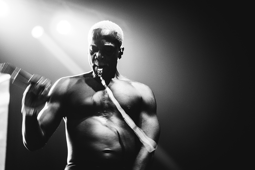 A shirtless performer energetically singing into a microphone on stage, with a whistle in his mouth. The performance is dramatically lit, emphasizing the intensity of the moment, indicative of a passionate live music show.