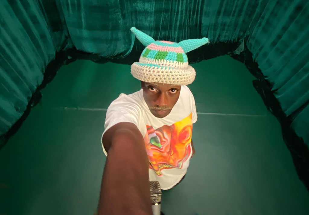 A person is taking a selfie from a high angle, capturing their upper body and face. They are wearing a whimsical, multicolored knitted hat with protruding horn-like features and a white T-shirt with a vivid, flame-like design. The background features what appears to be the textured, corrugated surface of a green tunnel, adding a sense of depth and a pop of color to the image.