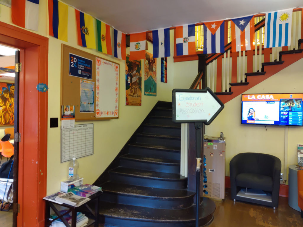 Interior entryway of La Casa Cultural Latina. The walls are a light yellow, with orange trim in the doorways and on stairs. The staircase goes up and turns to the right. There are flags hung along the walls, and lots of signage on bulletin boards and other wall space.