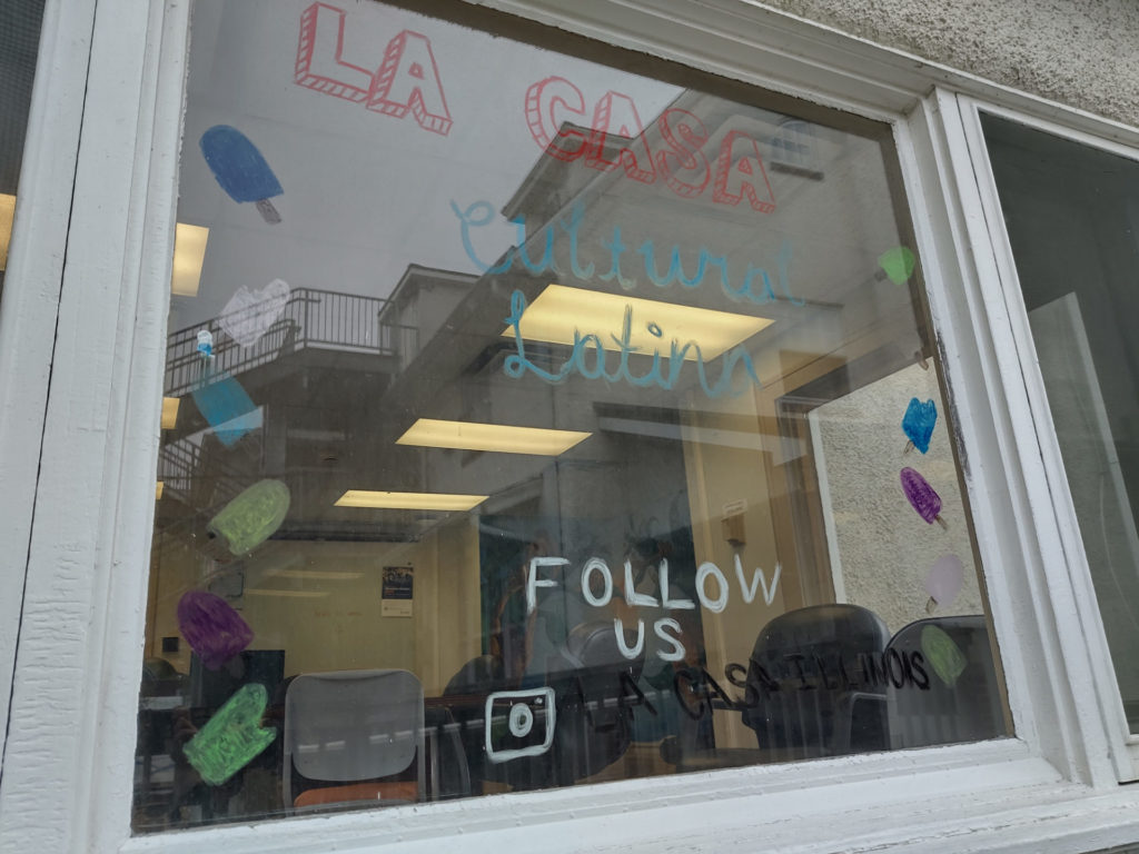 Window at La Casa Cultural Latina, with different writing on it that reads "La Casa Cultural Latina, follow us" with their Instagram handle below. Through the window you can see chairs around a table.