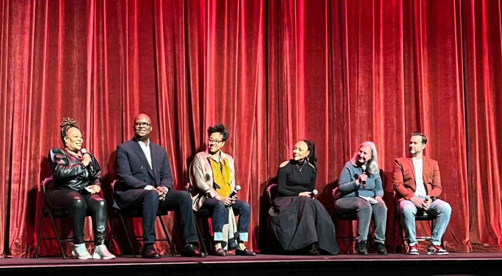 Six people sit in chairs on a stage with a velvet red curtain behind them. From left to right: panel moderator, a Black woman in an all-black leather outfit; Michael Swanson, a tall Black man in a suit; Christine Swanson, a Black-Korean American woman in a tan blouse and yellow scarf; Lynn Whitfield, a Black woman in an all-black dress; panel moderator, a white woman in jeans and a blue sweater; Brett Hayes, a white man in jeans and a red blazer.