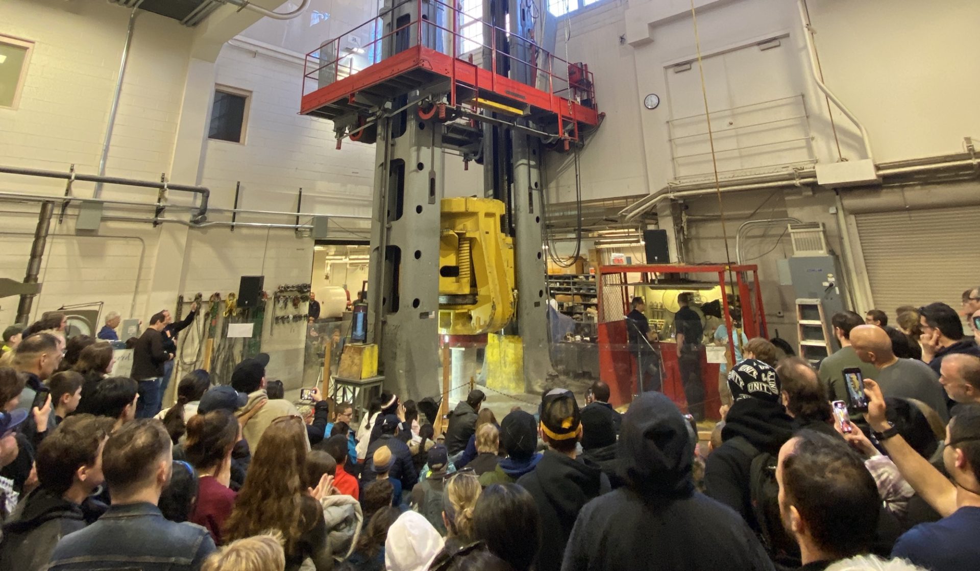 A concrete slab being crushed by a pressure machine two-stories tall, surrounded by a crowd of people