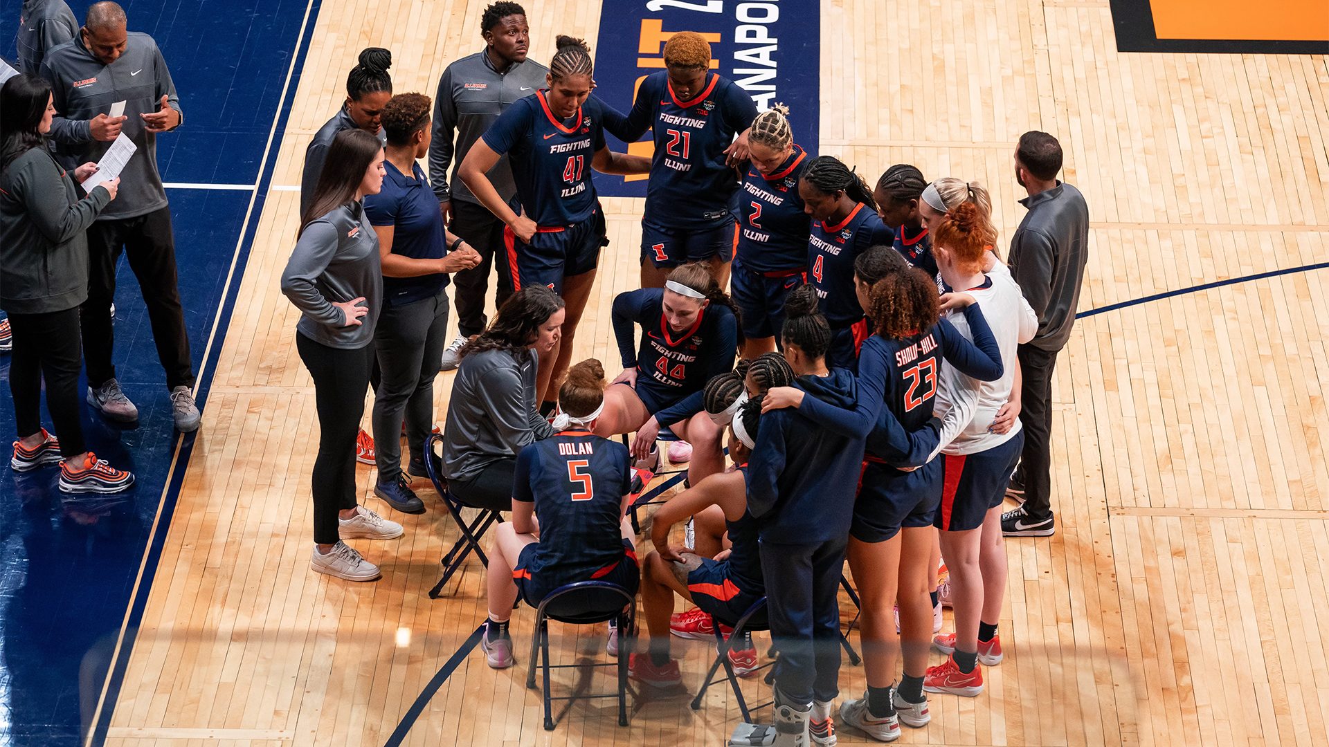 Players wearing dark blue jersey's with orange writing gather around their couch who is sitting on the basketball court.