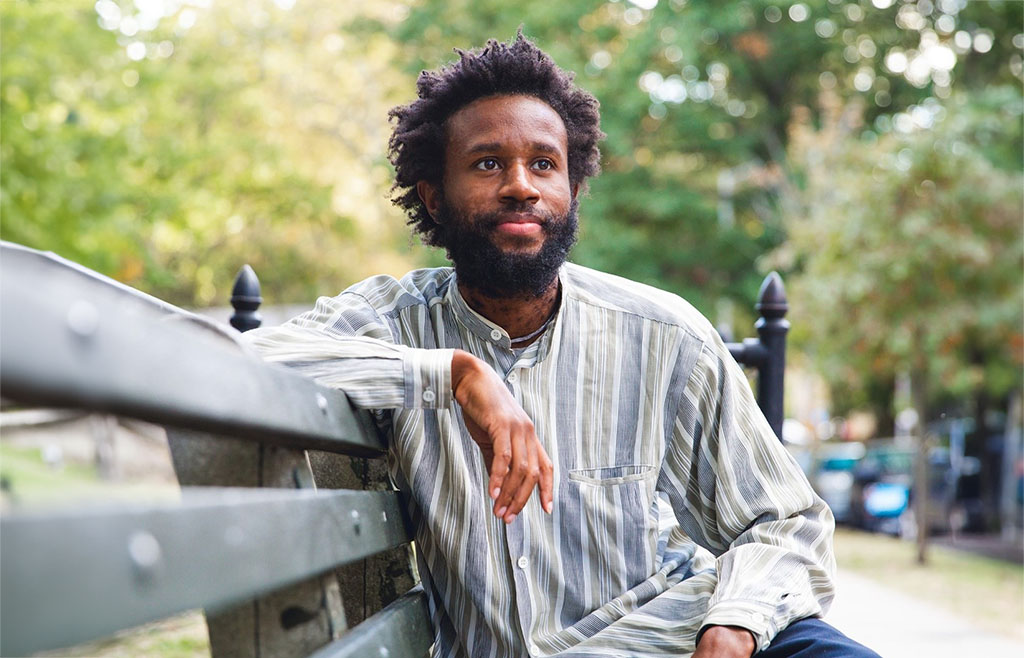 The image captures an individual sitting leisurely on a park bench. They are dressed in a casual, striped, button-up shirt with long sleeves, and they have a natural hair style. The person appears relaxed and reflective, with their arm resting on the back of the bench, and a gentle, thoughtful smile on their face. They are gazing off into the distance, possibly lost in thought. The background is filled with lush greenery, indicative of a park or a green urban space, and the blurred background suggests a peaceful, serene setting.