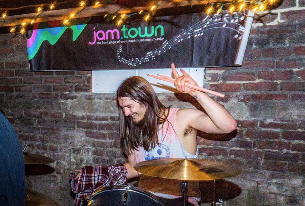 In a dimly lit, brick-walled room that speaks to the intimate gatherings of music enthusiasts, a drummer is captured in a moment of joyous release mid-performance. His long hair veils his face, which is adorned with a gratified smile, as he raises his drumsticks in a playful 'V' for victory or peace sign. He wears a light tank top, and the casual attire suggests a laid-back, yet impassioned musical event. In the background, twinkling lights add a warm ambiance to the space, while a banner emblazoned with "jam.town" in bright, bold letters hangs proudly, asserting the identity of the space as a local music hub. The banner's design, featuring an abstract wave and music notes flowing against a gradient of vibrant colors, encapsulates the essence of a community brought together by the universal language of rhythm and beat.