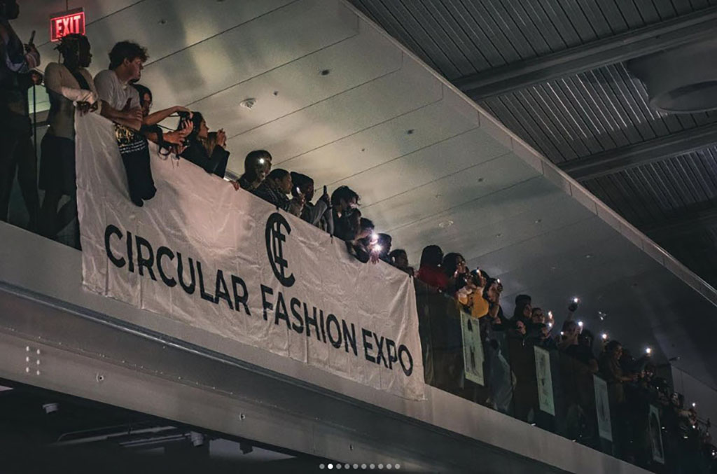 On an elevated indoor balcony, a lively brass band brings energy to an event. The musicians, in a diverse array of attire, signal a relaxed yet festive ambiance. They are arrayed along the railing, some with brass instruments at the ready, others clapping or capturing the moment on their phones. A banner prominently displayed over the railing reads "CIRCULAR FASHION EXPO," indicating the event’s theme focused on sustainable fashion practices. The soft, ambient lighting suggests the event is taking place in the evening, adding a sense of warmth and community to the gathering.