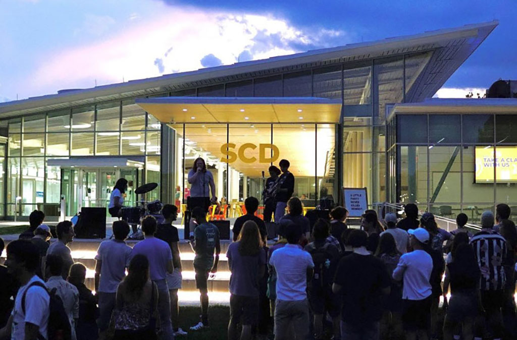 An outdoor live music event unfolds at dusk outside a modern building with large glass windows and an angled roof, creating a backdrop that reflects the evening sky. The audience, a diverse group of onlookers, is casually dressed, signifying a relaxed community event. They are focused on the performers on stage, with some members of the crowd sitting on steps that are illuminated from within, adding a soft glow to the twilight scene. The signage on the building includes the letters "SCD," possibly indicating the name or acronym of the venue. Musicians perform on a simple stage setup with a microphone stand and speakers visible, suggesting an intimate acoustic set. The entire scene is bathed in the serene light of the setting sun, casting a peaceful ambiance over the gathering.