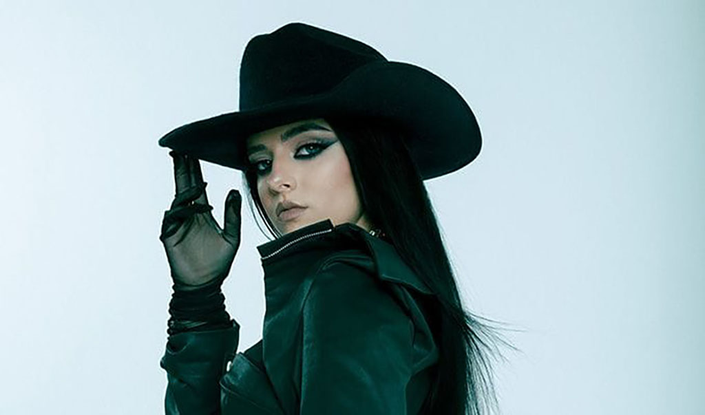 a person wearing a dark, wide-brimmed hat and a matching dark outfit with gloves. Their pose is striking, with one hand adjusting the brim of the hat, partially covering their face. The individual has dark hair and makeup that emphasizes their eyes. The background is a cool, solid color, giving the image a sleek, stylized look, indicative of a fashion-forward or dramatic persona.