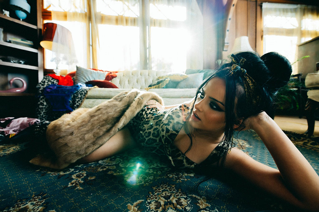 a person lying down on a patterned rug, propped on their elbow, surrounded by eclectic room decor that includes a mix of textiles and personal items. The individual is gazing at the camera with an introspective look, bathed in soft, natural light streaming in from windows. This casual, intimate setting suggests a candid or lifestyle portrait.