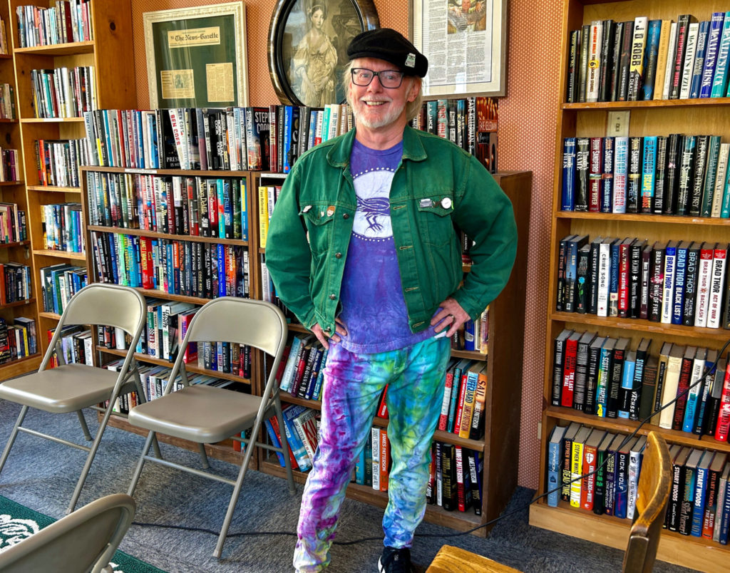 A colorful white man stands in front of large bookshelves. He is wearing a black cap, green jacket, purple shirt, and tie-dyed pants.