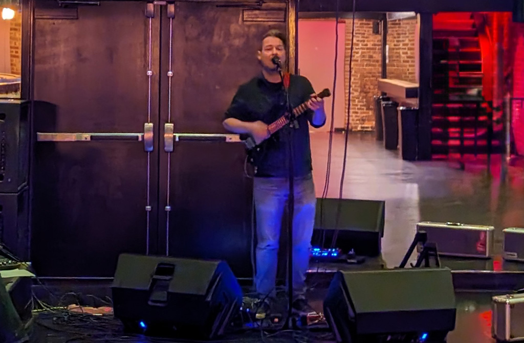 A musician stands on a stage, performing in front of a microphone. He's holding an electric guitar, intimately engaging with his small audience. The setting appears to be an indoor venue with mood lighting that casts a warm glow on the scene. Behind him, large, dark doors and brick walls suggest an urban, possibly industrial, venue typical for live music performances. Equipment cases are on the floor, indicative of a live event setup. The space beyond the performer is shadowy, with some ambient lights reflecting off the polished floor, hinting at the venue's spaciousness.