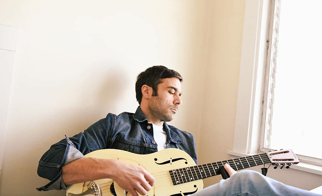 an individual seated while playing an acoustic guitar. The person is dressed casually in a buttoned denim shirt with rolled-up sleeves. The scene is peaceful and domestic, with natural light coming from a window to the right, creating a warm, inviting atmosphere. The person's eyes are closed, suggesting concentration or emotion connected to the music being played.
