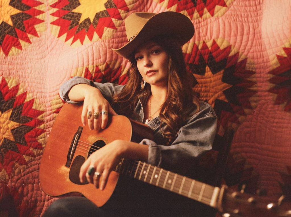 A girl with long brown hair wearing a cowboy hat and a denim jacket with a guitar in her lap looking at the camera.
