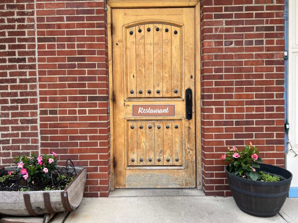 A wooden door with a simple brown sign reading "Restaurant" on a brick building with two planters of pink flowers.