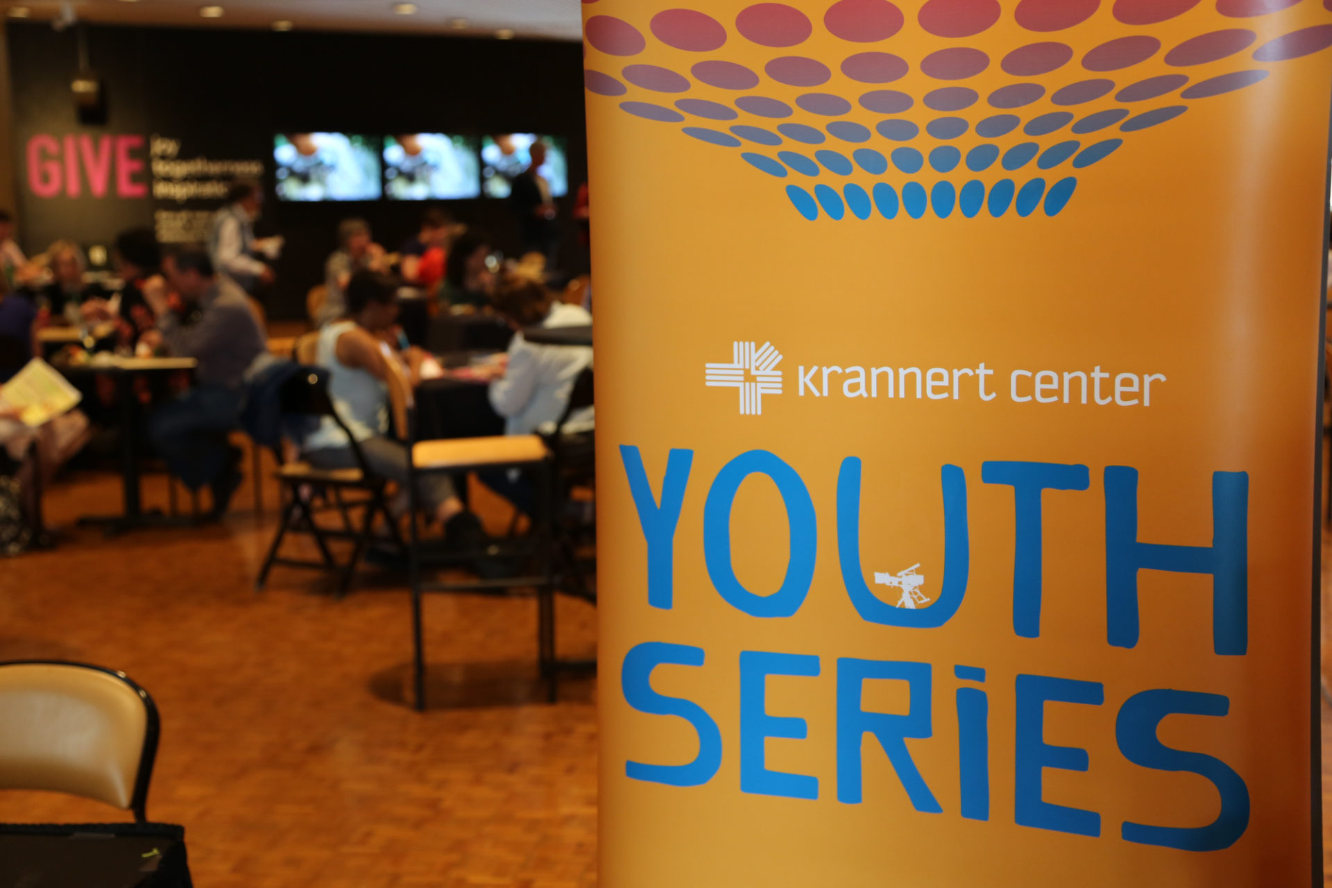 A yellow banner with the words "Krannert Center Youth Series" in white and blue text. The banner is in a room with a parquet wood floor, chairs, and tables. There is an out-of-focus accent wall in the back, with the word "give" in red or pink text on a black wall.