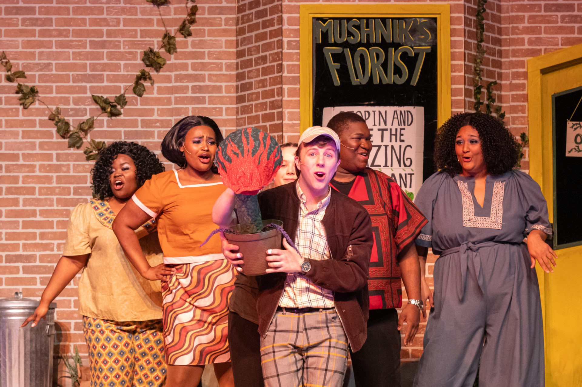 Photo of a white man holding a potted plant made of paper with a read flaming face on it. Behind him are four Black singers in beige, red, orange, and blue costumes on a stage set with the words Mushnik's Florist on a sign.
