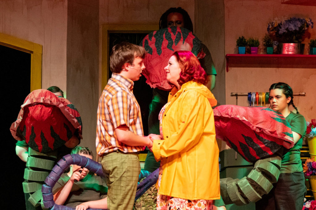 A white man and a white woman face each other on a stage. Behind them are the red and green puppet pieces manipulated by three people holding parts of the puppet.