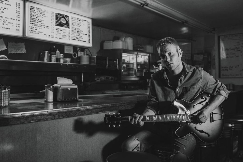 A black and white photo with a man with short-cropped hair wearing a denim shirt playing guitar while sitting down at a diner.