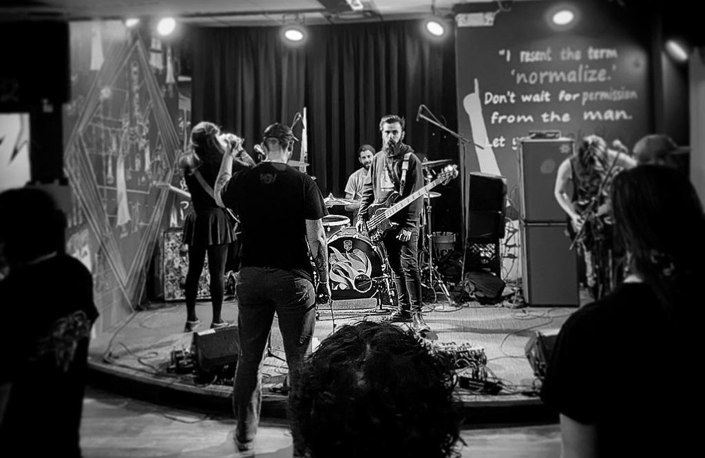The venue is intimate, featuring vibrant murals and a political banner that reads, "I resent the term 'normalize.' Don't wait for permission from the man. Let your freak flag fly." A four-member band performs energetically on stage, engaging with the audience. One band member, central to the scene, sports a hoodie and plays a black bass guitar. A vocalist, possibly a woman based on attire, wears a black outfit and uses a microphone. Another guitarist, with a beard, wears a casual shirt and plays an electric guitar. The drummer in the background, partially obscured, contributes to the dynamic scene.