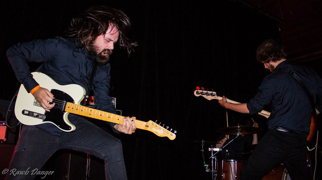 In this dynamic photo, two musicians are captured mid-performance, passionately immersed in their craft. The musician on the left, with long hair obscuring part of his face, strikes an intense pose as he plays a white electric guitar, his body leaned forward in a moment of fervor. On the right, another musician is bent over a drum set, focusing intently as he plays the bass guitar, both members dressed in dark attire that reflects the serious and energetic atmosphere of their performance. They are onstage, illuminated against a dark background that contrasts with the vitality and movement suggested by their postures and expressions.