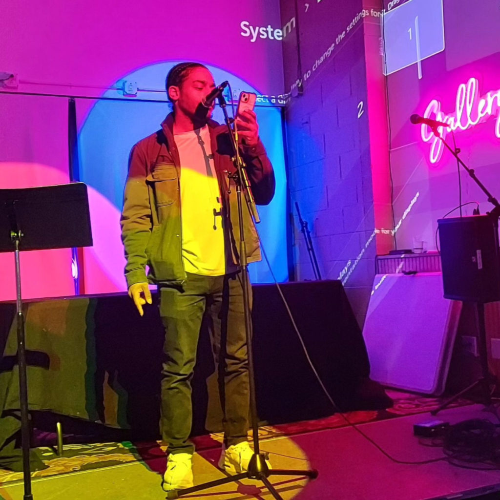 A Black man stands at a microphone reading poetry from his mobile phone. The stage is colorfully lit with bright pink and blue lights.