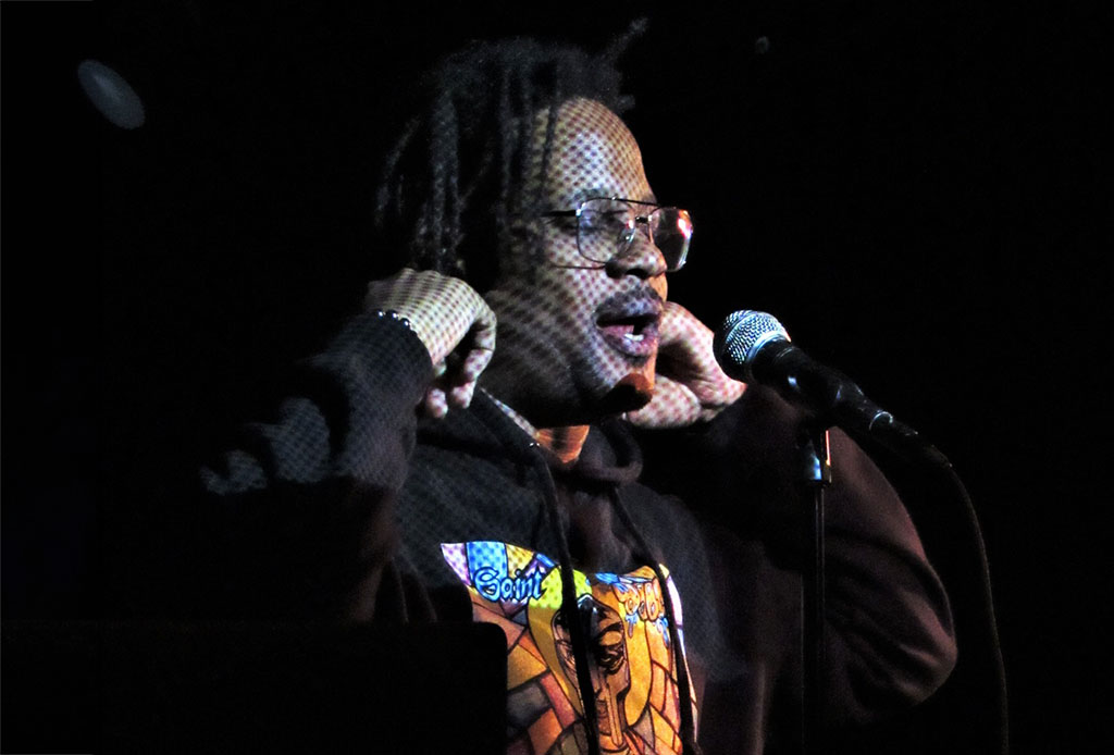a performer on stage, in the act of speaking or singing into a microphone. The performer is wearing a dark outfit with a checkered sleeve and glasses, lit by the contrasting stage lighting that creates a dramatic effect. This moment on stage suggests a live performance, perhaps of a musical or spoken word nature, to an unseen audience in a dark venue.