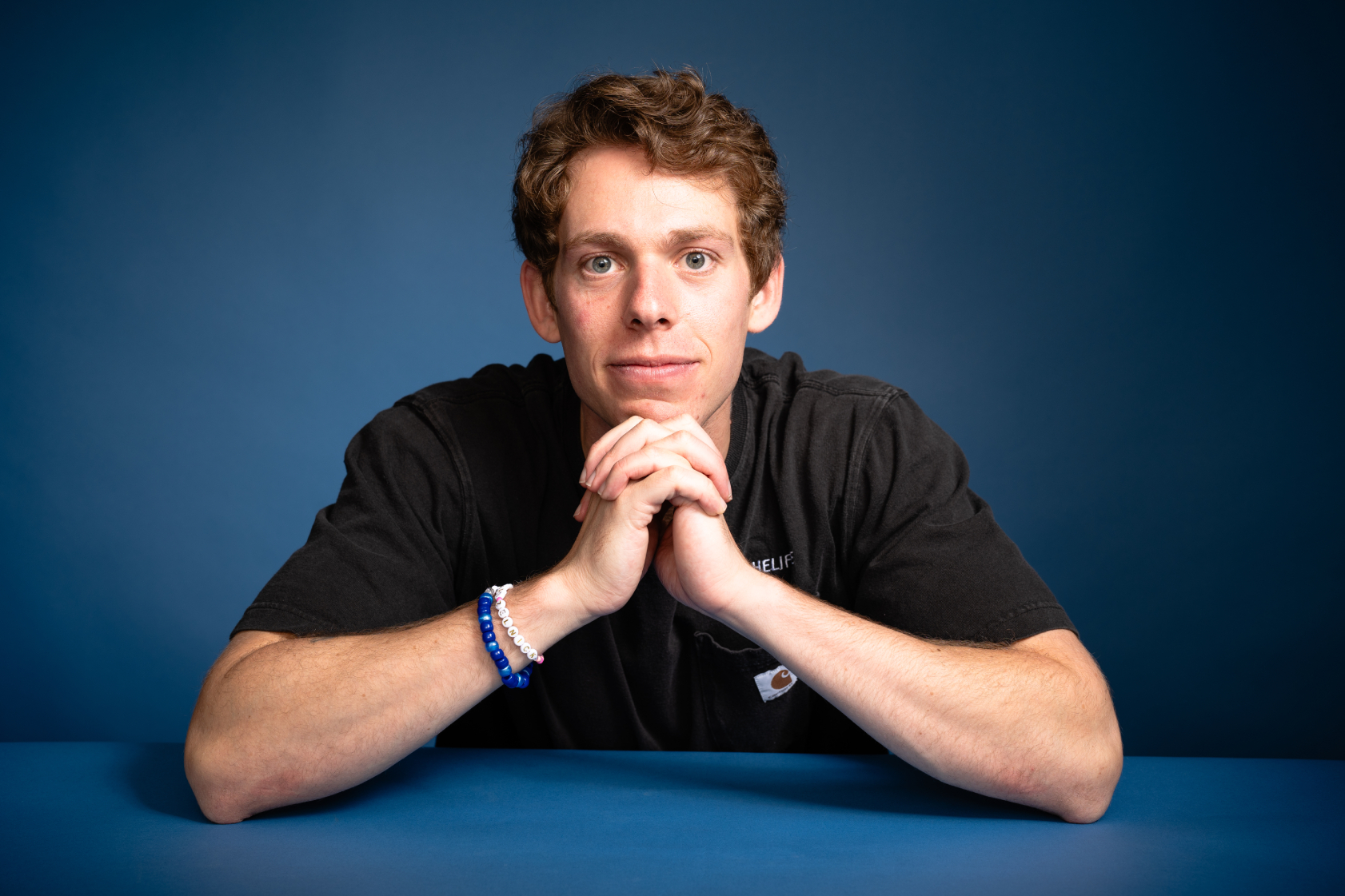Portrait of Lucas Zelnick, a white man in his 20s, wearing a black t-shirt. His hands are folded under his chin. The backround and table on which he is leaning are blue. He has light brown hair.