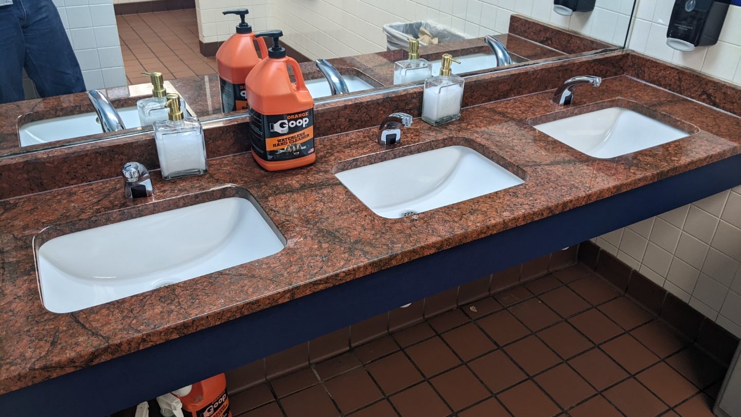 A bathroom with three white sinks on an orange tile counter. There is a large mirror and on the sink is a large plastic bottle of cleaner. 