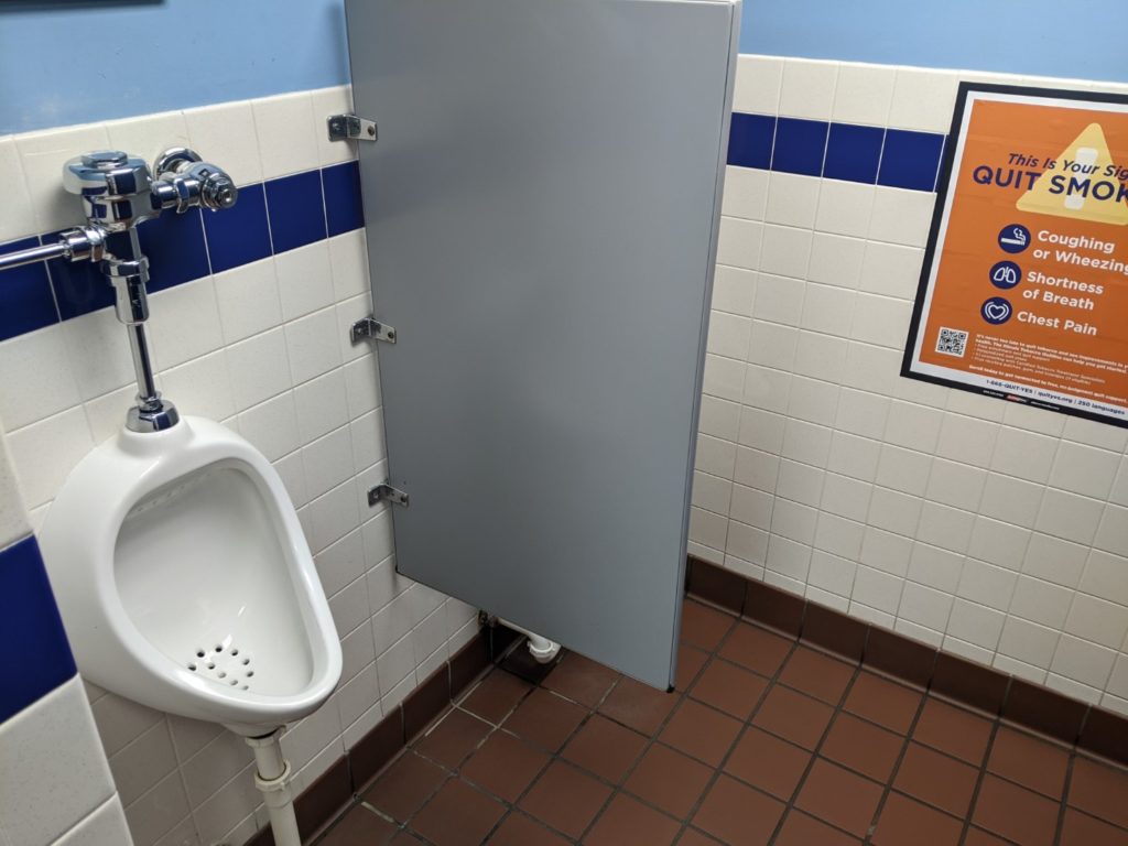 A bathroom with a brown tile floor, and light blue walls with an orange sign that says "quit smoking". There is a white urinal next to a gray dividing wall. 