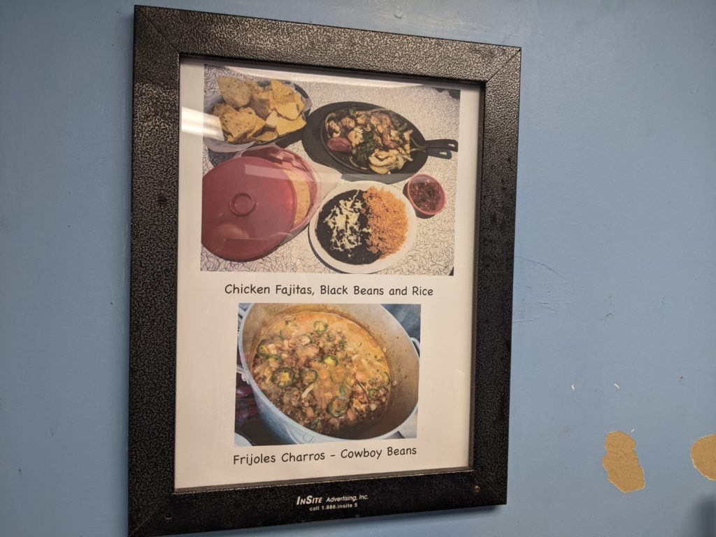 A black framed menu items. One is for chicken fajitas, black beans and rice. The other for frijoles charros- cowboy beans. The wall is light blue. 
