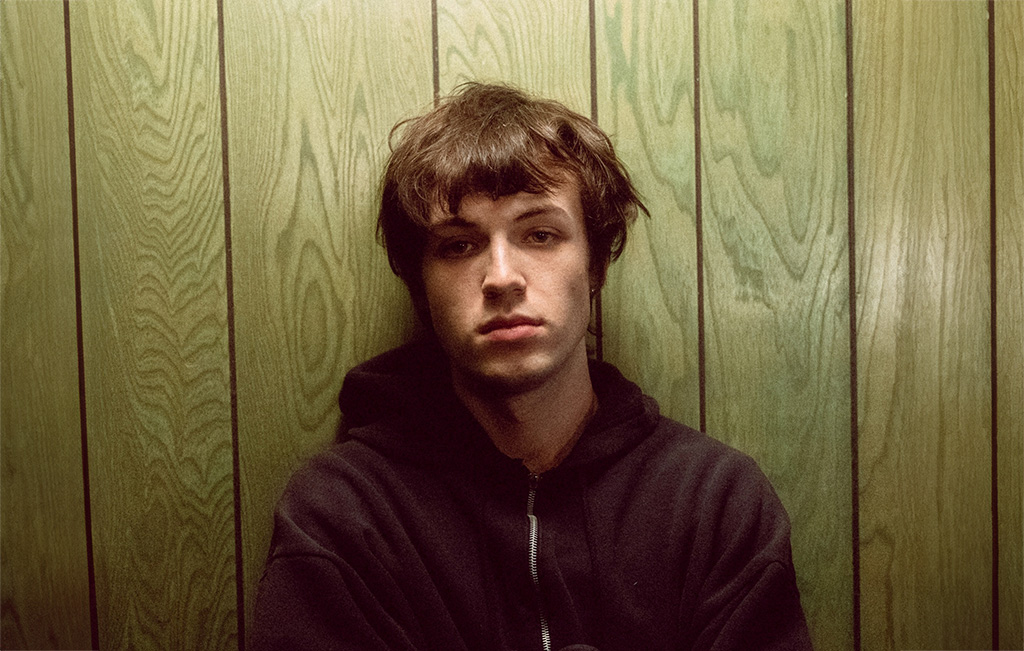 an individual with a contemplative or somber expression, captured in a room with wooden paneling in the background. The person is wearing a dark hoodie and their hair is a shaggy, chestnut brown, partially covering their forehead. The overall tone of the photo is quite subdued and reflective, with a sense of introspection or pensiveness.