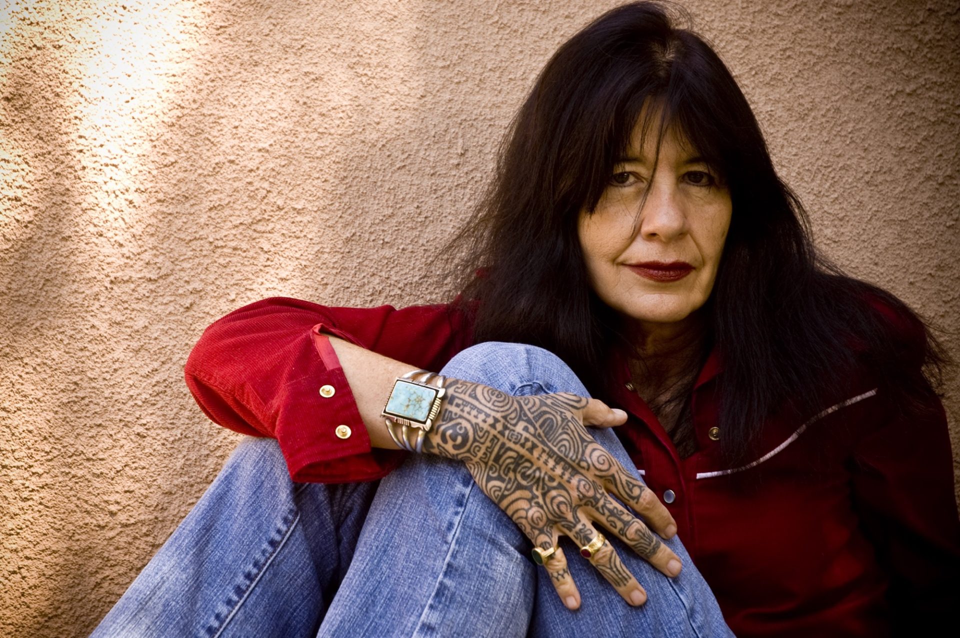 Native American woman sitting wearing a red shirt and blue jeans, her tattooed hand placed on her knee