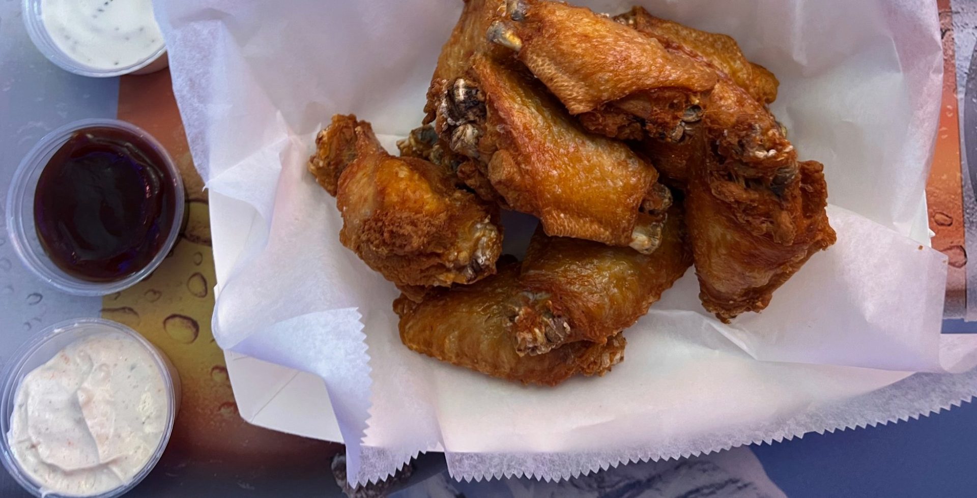 An order of ten wings in a parchment-paper lined basket beside two sauces (one ranch, one barbecue, and one white zesty sauce).