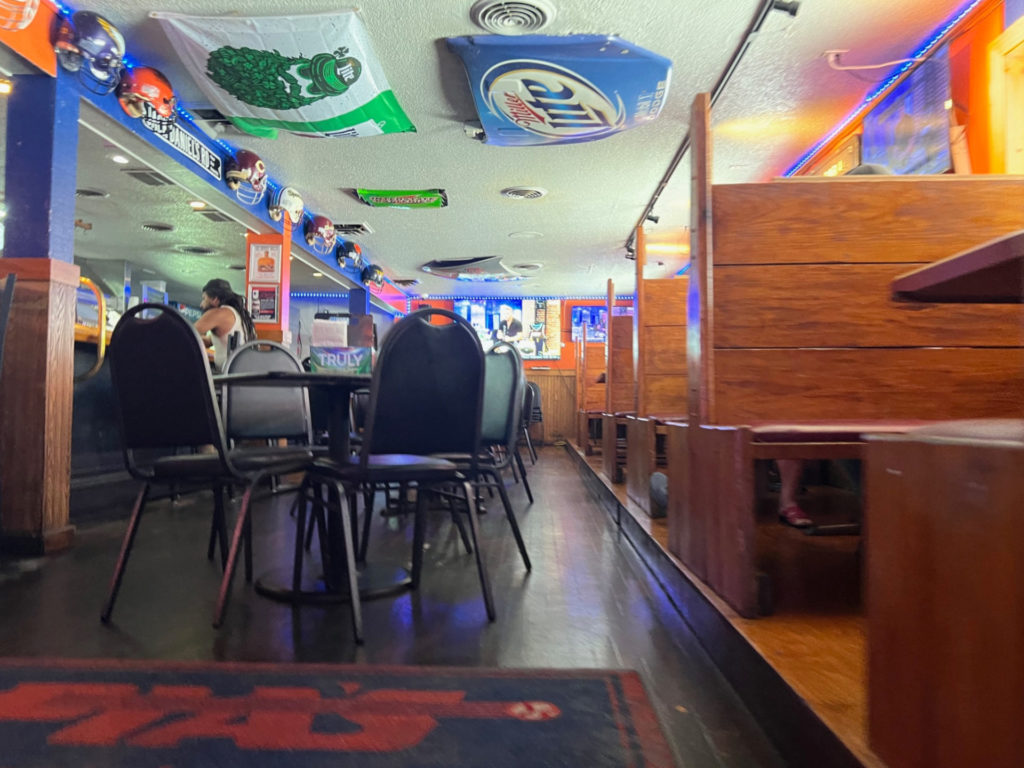 The interior of Pia's Sports Bar has wooden booths, black tables, and bar seating long a blue bar with football helmets from different teams hanging above it.