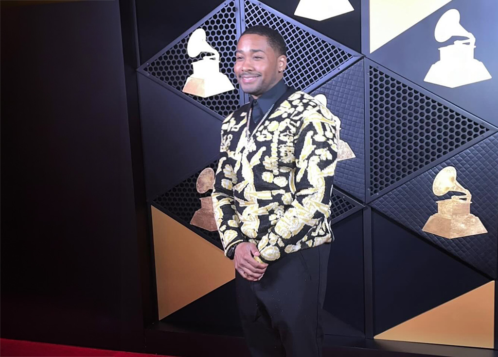 A young man dressed in a gold and black jacket with black pants stands in front of a backdrop to get his photo taken at the Grammy Awards.