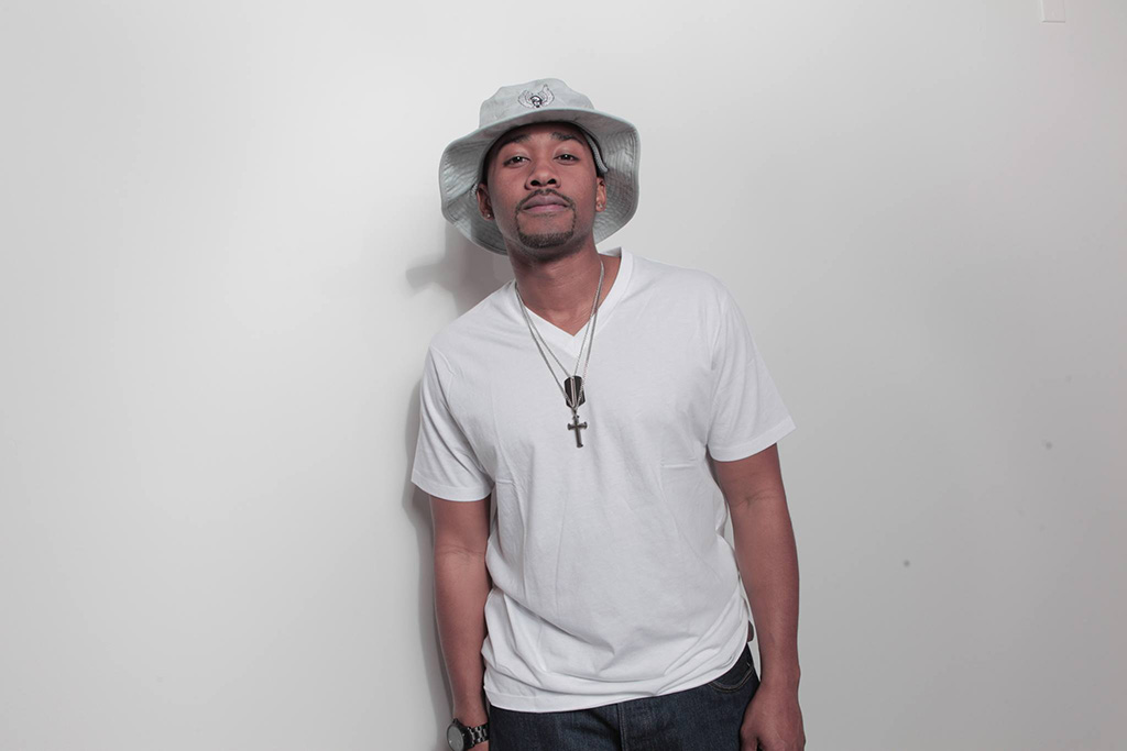 A man in a white t-shirt and white bucket hat leans back on an off-white wall with his hands in his pockets.