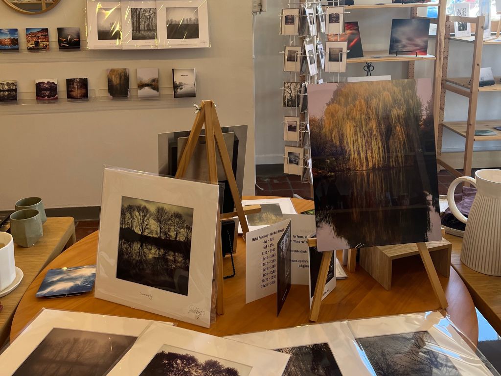 Artwork can be seen on the foreground of a wooden table and on the wall behind is shelves with more pictures in them, and a spinning rack with postcard sized images.